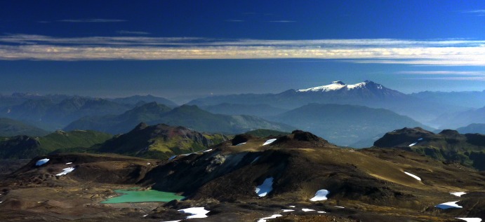 A sub-ice fissure eruption formed the ridge above the green lake, with lava sheets and lobes flowing beneath the ice away from the ridge. The distinct snow-capped twin peaks of Mochu-Choshuenco volcano are in the background.